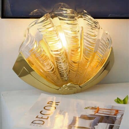 on a white table with a decoration magazine on it, we see a brass and glass wall sconce with a transparent shade, the sconce is shaped like a shell
