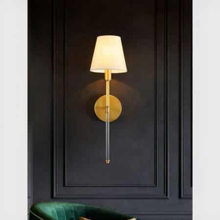 Brass wall light with classic fabric shade, lit and mounted on a gray wall.
