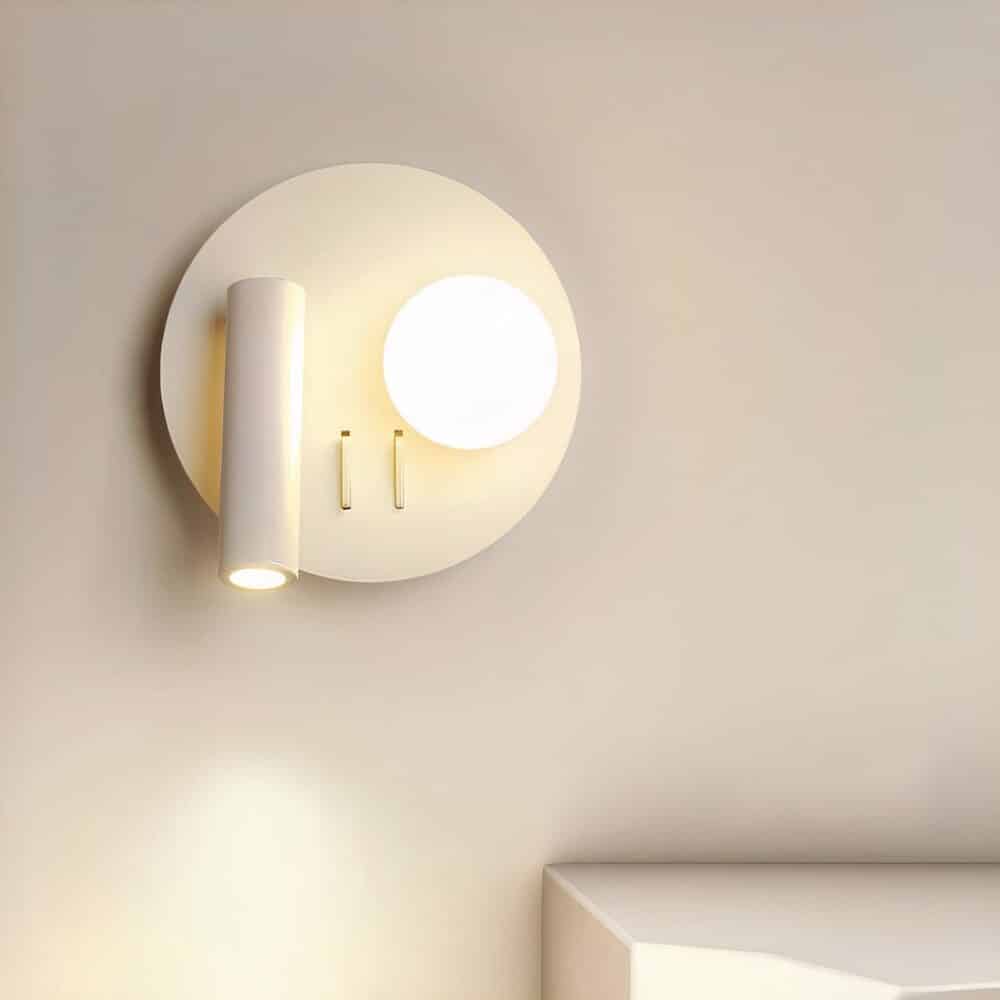 Round white wall lamp with 2 light sources: on the left, a tube-shaped reading lamp shines downwards, and on the right, a lighted ball. Mounted on a white wall.