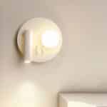 Round white wall lamp with 2 light sources: on the left, a tube-shaped reading lamp shines downwards, and on the right, a lighted ball. Mounted on a white wall.