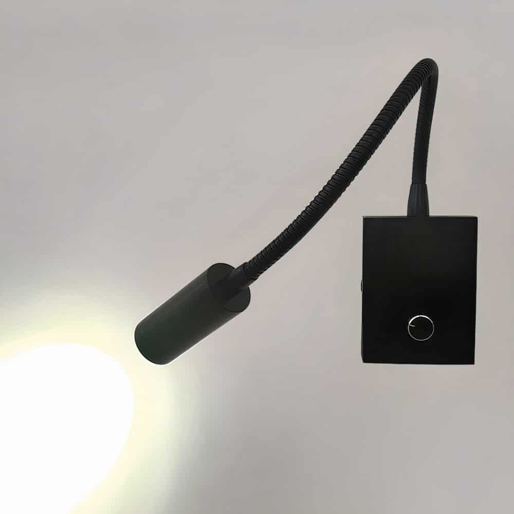 Black wall reading lamp with rectangular base with knob, flexible stem and light head that shines a cool light towards the wall. The wall is gray.