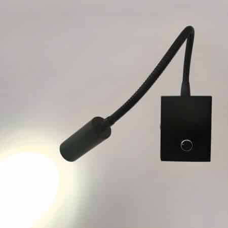 Black wall reading lamp with rectangular base with knob, flexible stem and light head that shines a cool light towards the wall. The wall is gray.