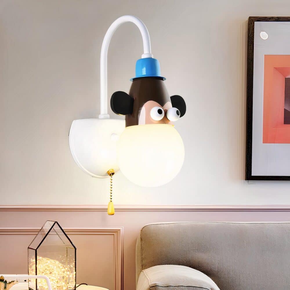 White wall sconce for children's room with half-ball wall bracket and angled rod supporting a monkey head up to its eyes and a luminous ball below. The wall light is fitted with a gold chain. Attached to the wall is a picture frame on the right, a light-brown sofa underneath and objects to the left.