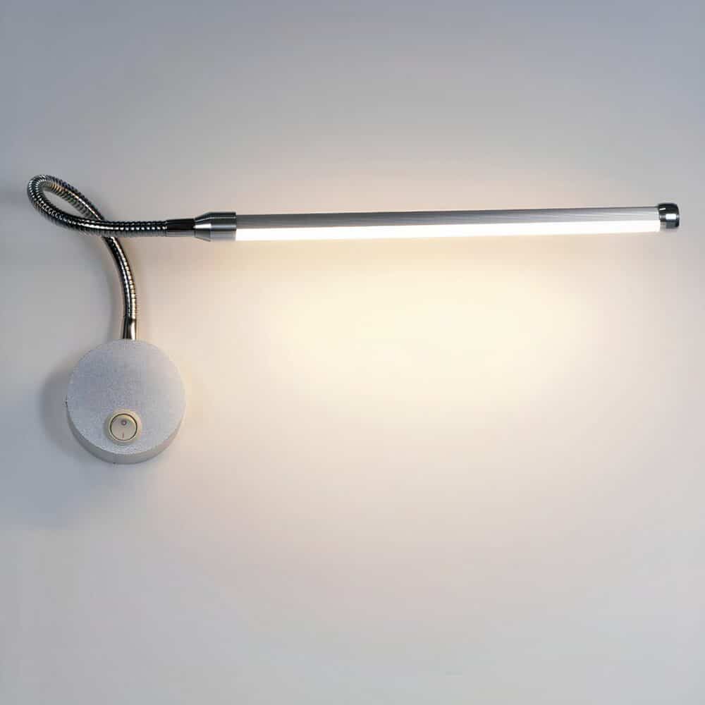 Grey wall-mounted reading light with round base and switch, flexible stem and long light head. On a white background, with the reading light as the only light.