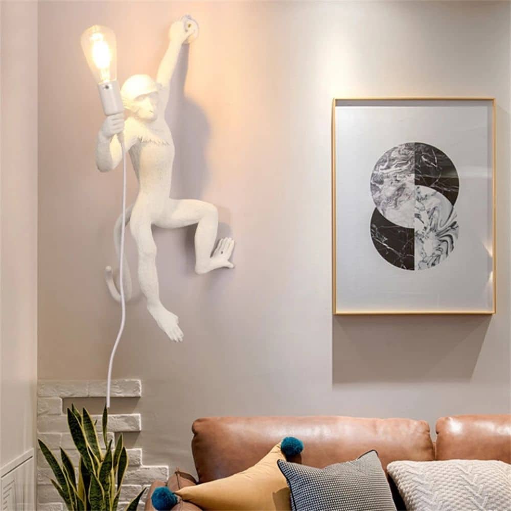 Wall light in the shape of a monkey hanging on the left side in a white version and hung on a white wall in a living room, above a sofa and a green plant.