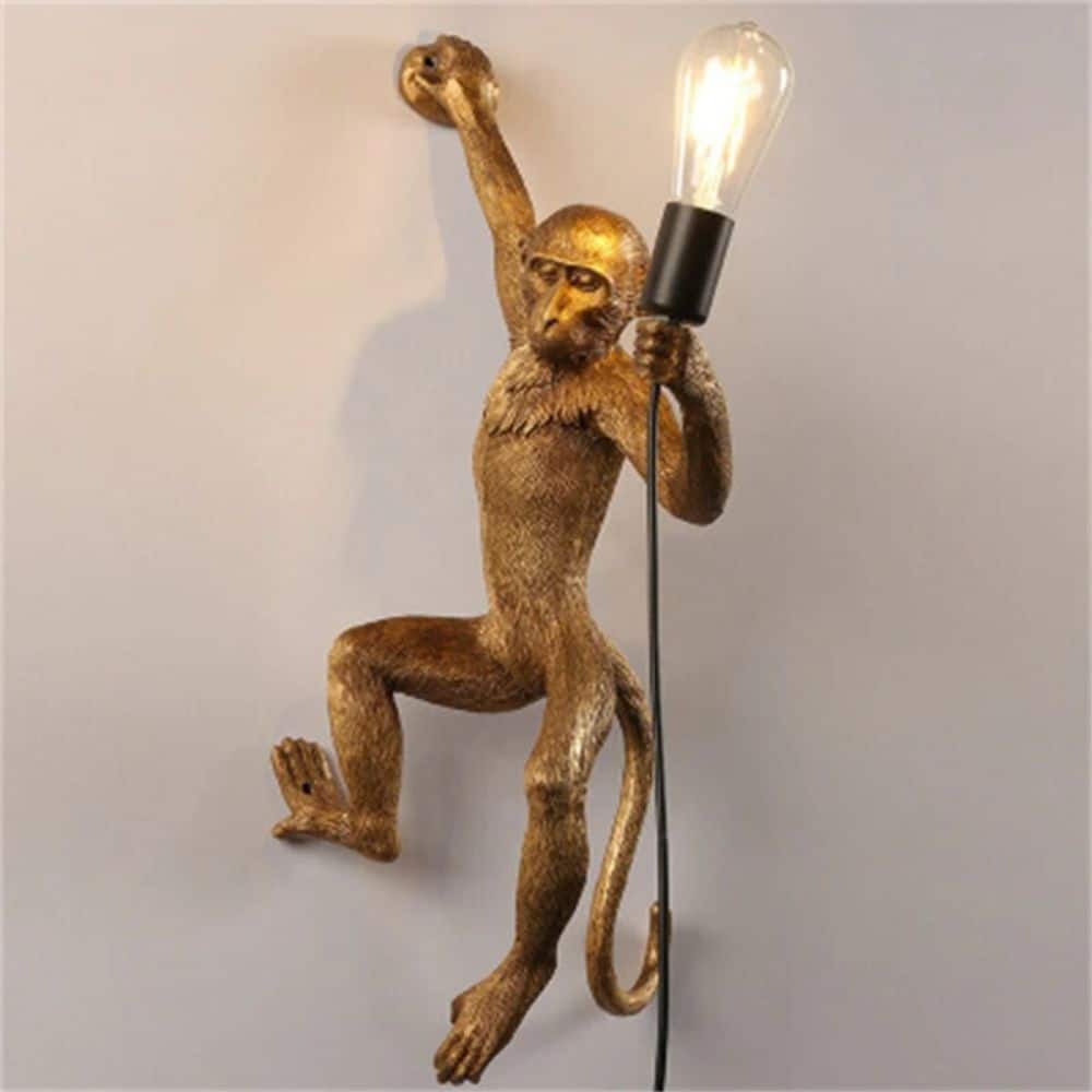 Wall light in the shape of a monkey hanging on the right-hand side, in gold version and hung on a white wall.