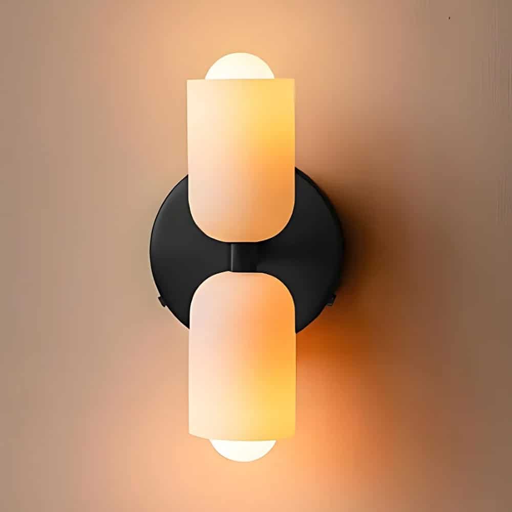 Wall sconce with double head up and down. The heads are white cylindrical with one bulb lit. The round base fixed to the wall is black.