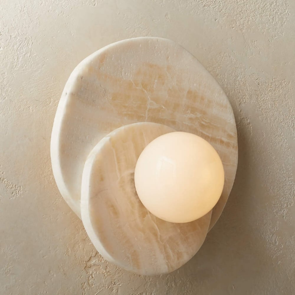 Circular wall light in natural stone composed of two beige stone rounds, the smaller on the larger with a luminous ball in the center. Mounted on a beige wall.