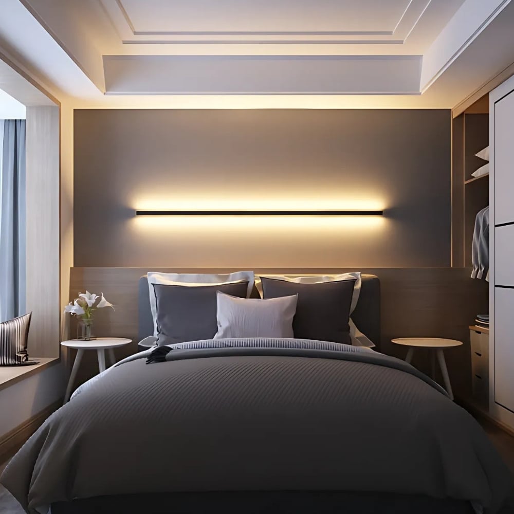 Black minimalist wall light in the form of a horizontally fixed tube with warm light above and below. This wall light is mounted in a bedroom above a double bed with gray bedding.