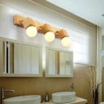 Wooden cube-shaped bathroom wall light with 3 light sources. The bulbs are switched on. The wall light is fixed above two rectangular mirrors, themselves above two white washbasins.